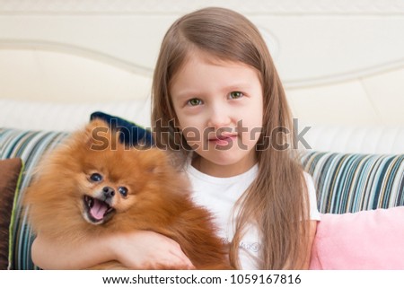 A pretty little girl with brown hair is reading a book and watching cartoons on her computer with her dog. Friendship. Pomeranian Spitz.