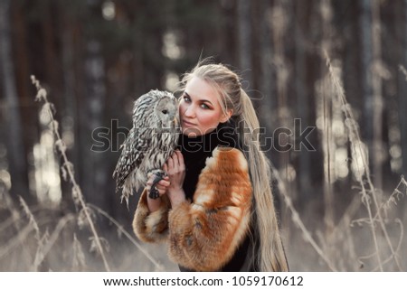 Blonde woman with an owl in her hands walks in the woods in autumn and spring. Long hair girl, romantic portrait with owl. Art fashion photo, beautiful makeup