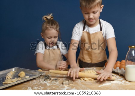 Picture of blonde 7 year old boy with hair knot helping his elderly brother to bake cookies at kitchen counter. Two preschoolers making dough for pasty, using rolling pin, milk and eggs on table