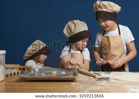 Picture of confident schoolboy in apron and chef hat teaching his little brother and sister to make ginger cookies, holding metal mold, explaining them how to use it right. Family and baking