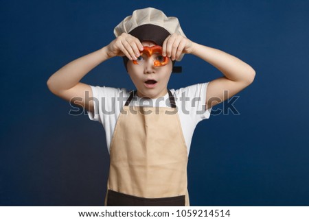 Children, fun and cooking. Picture of astonished 10 year old European male kid in apron and cap posing in studio with slice of red bell pepper, having shocked look, keeping mouth wide opened