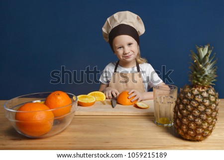Picture of cute blonde 7 year old boy in chef uniform standing at wooden kitchen table and slicing orange while learning how to make fruit salad at cooking workshop for children, smiling happily