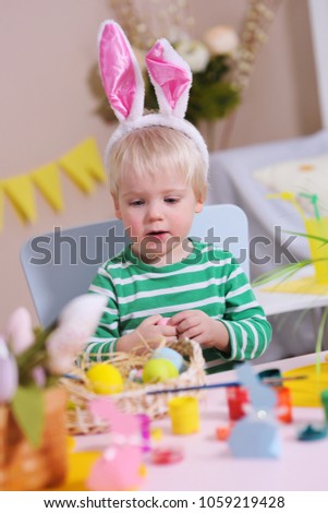little cute baby boy with white hair with rabbit ears on his head sitting at a table in front of an Easter basket and decor. Preparation for the celebration of Easter
