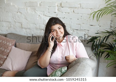 People, modern technologies and communication concept. Picture of beautiful overweight happy young brunette woman wearing pink shirt smiling happily while talking on mobile phone with her boyfriend