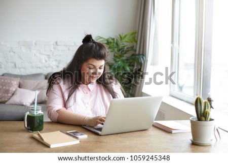 Picture of beautiful chubby young female journalist with hair bun typing article for women's beauty magazine about body positive movement, having green fresh juice. People and modern technologies