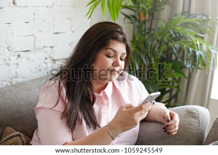 Indoor shot of overweight young dark haired female in pink shirt having disappointed look while texting sms using mobile phone at home. Brunette chubby girl messaging friends online via messenger app