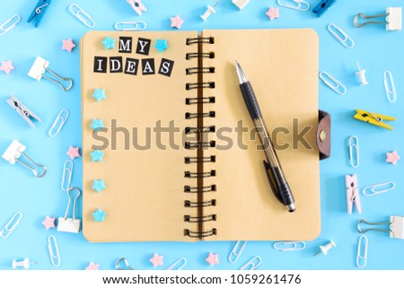 Notepad on springs with an inscription My Ideas. Chaotically scattered office supplies b and small asterisks on a blue background. Beautiful picture from above.