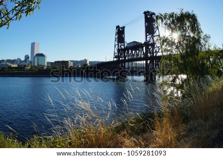 View of Steel Bridge in Portland Oregon with Buildings, Water, Trees, Grass, and Sun