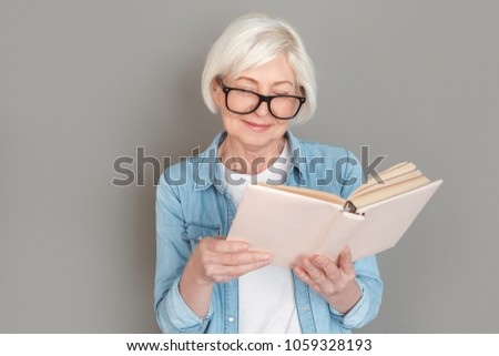 Senior woman in jeans jacket studio isolated on grey wall reading book joyful