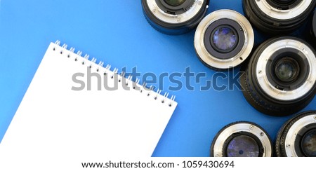 Several photographic lenses and white notebook lie on a bright blue background. Space for text