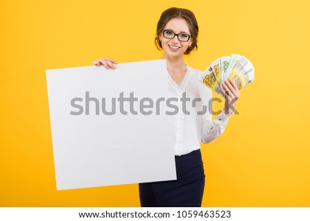 Portrait of confident beautiful young business woman with money in her hands and blank billboard on yellow background