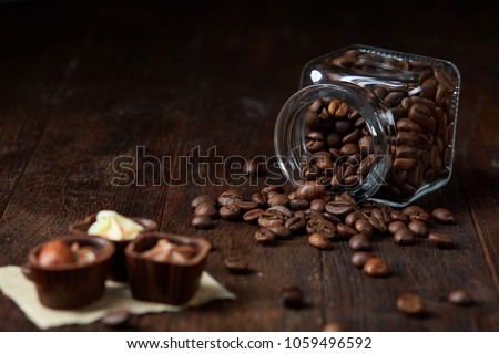 Side view of overturned glass jar with coffee beans and chocolate candies on wooden background, selective focus