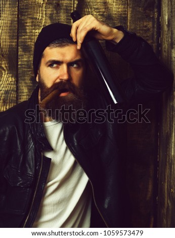 Bearded man, long beard. Brutal caucasian serious unshaven hipster holding bottle and smoking cigar with black leather jacket, hat and white shirt on brown vintage wooden studio background