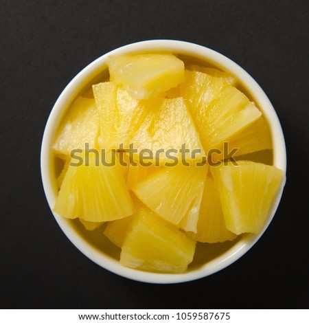 Pieces of pineapple cut into slices in a white bowl. Ripe yellow sweet fruit on black background. Royalty-Free Stock Photo #1059587675