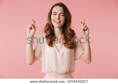 Portrait of a smiling young girl in dress holding fingers crossed for good luck isolated over pink background
