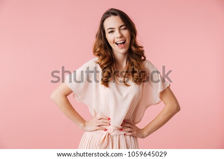 Portrait of a cheerful young girl in dress looking at camera and winking isolated over pink background