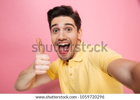 Portrait of a happy young man showing thumbs up gesture while taking a selfie isolated over pink background