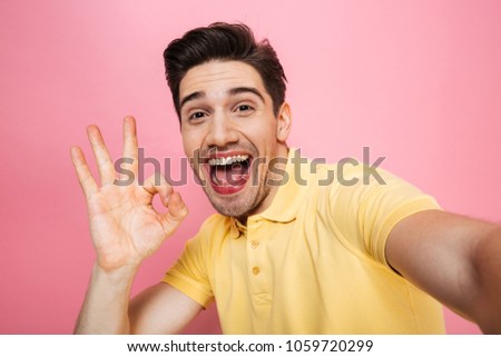 Portrait of a happy young man showing ok gesture while taking a selfie isolated over pink background