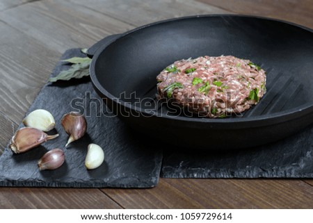 A piece of raw minced meat with herbs and garlic on a cast iron grill pan lies on a gray stone.