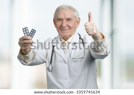 Senior therapist with two packs of medicine. Portrait of a confident old male doctor with stethoscope dressed in uniform holding pills pack and showing thumbs up gesture.