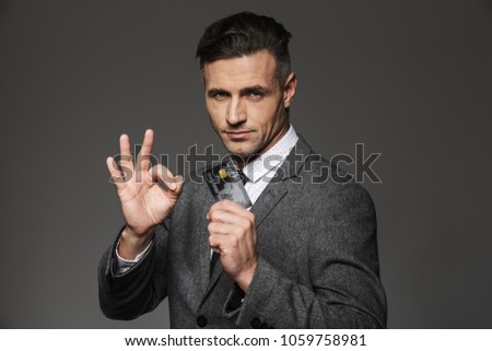 Rich and smart man 30s in formal suit and tie demonstrating plastic credit card and gesturing ok sign isolated over gray background
