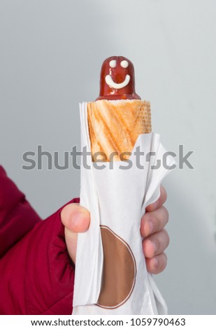 The hand holds hot dog with a smile and ketchup in a close-up on a background a white wall