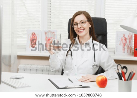 Female doctor sitting at desk, working on computer with medical documents in light office in hospital. Woman in medical gown with apple, glass of water in consulting room. Healthcare medicine concept