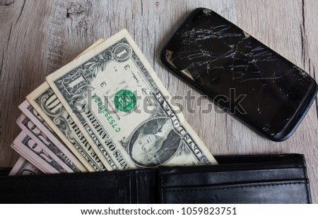 Crashed black smartphone and money in leather wallet to buy a new one. Dollars and broken phone on the woodeh background. Time to get a loan for electronics.