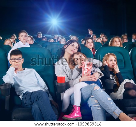 Pretty senior sister is watching movie or cartoon in the new cinema hall with junior children . children looking very interested and exited. There are other people on the backgroung.