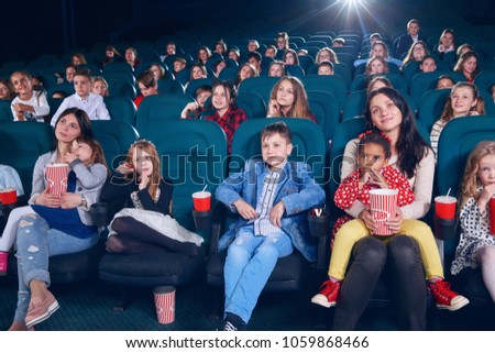 Mother watching intresting movie with children on the first row of the cinema hall. Kids are emotional,exited and satisfied. They smiling, laughing, drinking fizzy drinks and eating popcorn.