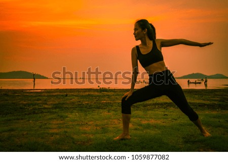 A slim young woman pratices yoga on the lake shore during a vibrant sunset