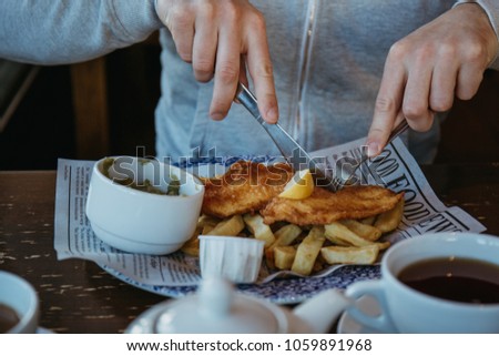 Man sitting at the wooden table, eating fish and chips, traditional English dish, tea and sauces near. Royalty-Free Stock Photo #1059891968