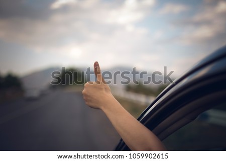 Young happy lady driving a car and making Like / Ok sign with hand out of window. Girl driving and enjoying road trip and travel, toned with soft autumn colors.