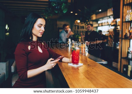 Nice picture of beautiful girl sitting at the barman's stand in the club and looking to the phone. She puts her left hand on the glass with cocktail. Barman and another guy are looking to her.