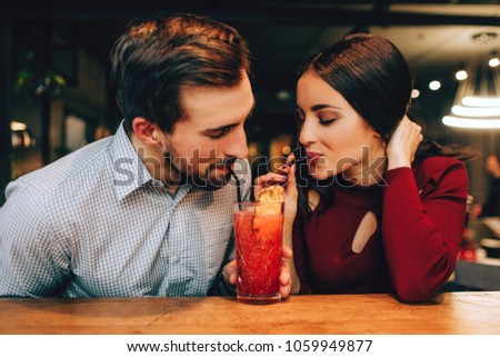 Nice picture of young couple sitting together and drinking red cocktail from the same glass at the same time. They look happy together.