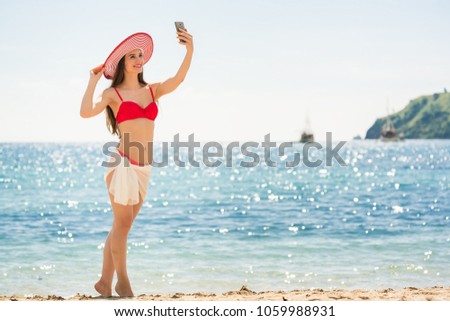 Beautiful young woman wearing a trendy striped hat while posing for a selfie picture on the beach during summer vacation in Flores Island, Indonesia