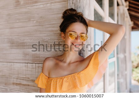 Portrait of laughing ecstatic lady with brown hair enjoying vacation at resort. Photo of inspired tanned woman expressing happiness while standing near wooden house.