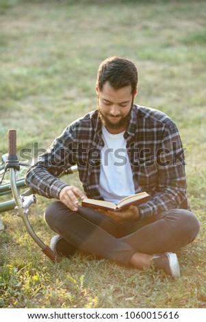 Handsome bearded man reading interesting book, enjoying the leisure while sitting on the grass near the bike in the park, wearing casual clothes.