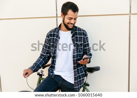 Smiling stylish man using a telephone, chatting, browsing websites, while sitting on bicycle, having rest at sunny day, outdoors.