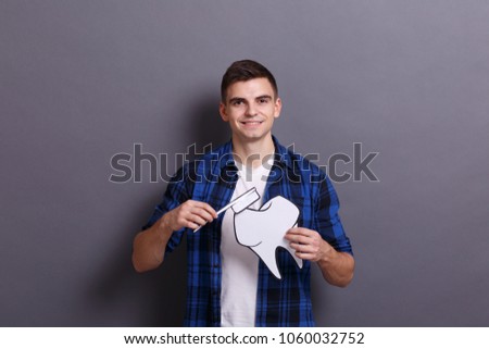 A young man holds a paper image of a tooth and a toothbrush on a gray background. Healthcare concept.