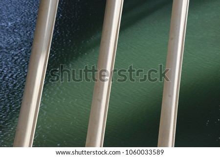 Close up outdoor view of many posts under a bridge desk. Abstract image composed of pillars pattern with a dark green river in background. Parallel grey iron tubes in front of the water surface. 