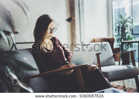 Young beautiful blonde woman sitting at the comfortable armchair and using laptop computer.Working process at coworking studio.Panoramic windows on blurred background
