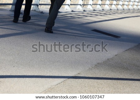 View of feet of people going away on a bridge. Leading lines with a metallic barrier and its shadow on the sidewalk. Graphic image with legs and rails. Grey ground surface with long dark shadows. 