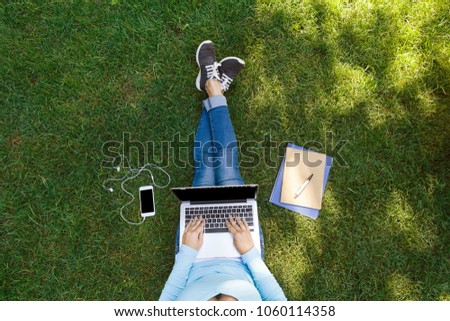 Top view of girl sitting in park on the green grass with laptop, notebook and smartphone with earphones, hands on keyboard. Copy space for text
