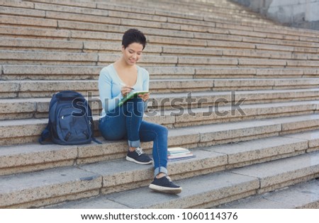 Preparing to exams outdoors. Beautiful young female student writing something in notepad sitting on the steps. Education concept, copy space