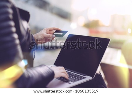 Cropped photo of a girl in black biker leather jacket sitting on a bench outdoors with her laptop, holding credit card in hand and making online purchases. Internet shopping concept. Close up.