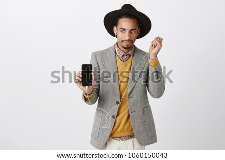 What is wrong with sound. Surprised worried handsome guy in trendy clothes and hat, showing smartphone to camera, taking off earphone and staring at telephone with anxious, tensed expression