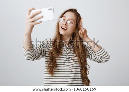 Popular fashion blogger makes new vlog using smartphone. Portrait of positive confident european girl showing v sign near face, making cool smiling expression while taking selfie over gray background