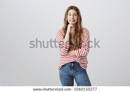 Yikes, mom saw broken vase. Studio shot of pretty embarrassed teenage girl holding hand on chin and grimacing with awkward expression, being guilty and hoping that nobody will know about her fault Royalty-Free Stock Photo #1060150277