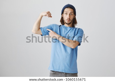 Do not underestimate skater boy. Portrait of attractive funny young guy in beanie and blue t-shirt raising arm and pointing at muscle, showing his strength, ensuring that he can protect girlfriend